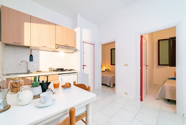 Cucina luminosa con tavolo apparecchiato. Due stanze sullo sfondo con porte rosse.