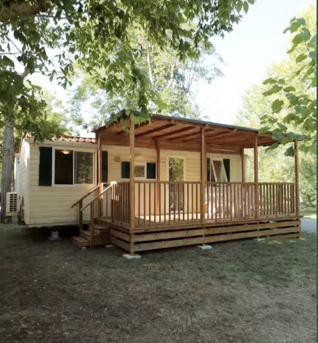 Una casa mobile con veranda in legno chiaro situata tra alberi verdi.