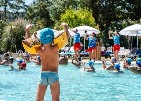 Bambini con braccioli gialli e cuffie azzurre in piscina, mentre istruttori guidano un'attività.