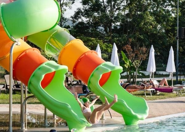 Persona su uno scivolo acquatico verde a spirale che scende in piscina. Ombrelloni sullo sfondo.