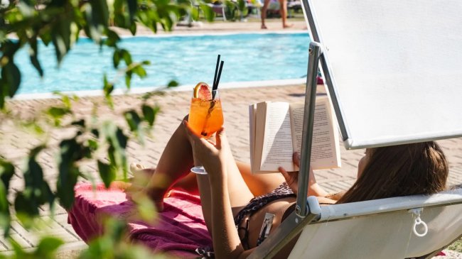 Una persona su un lettino, con un libro e un cocktail arancione in mano, accanto a una piscina.