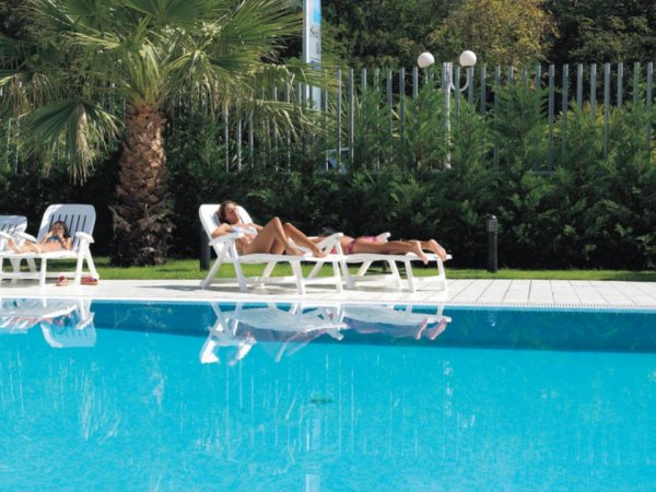 Persone sdraiate su lettini a bordo piscina, tra palme.