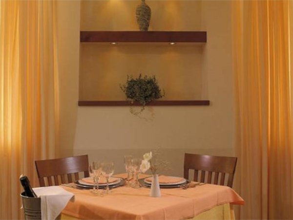 Tavolo apparecchiato per due in una sala con scaffali decorativi e tende arancioni.
