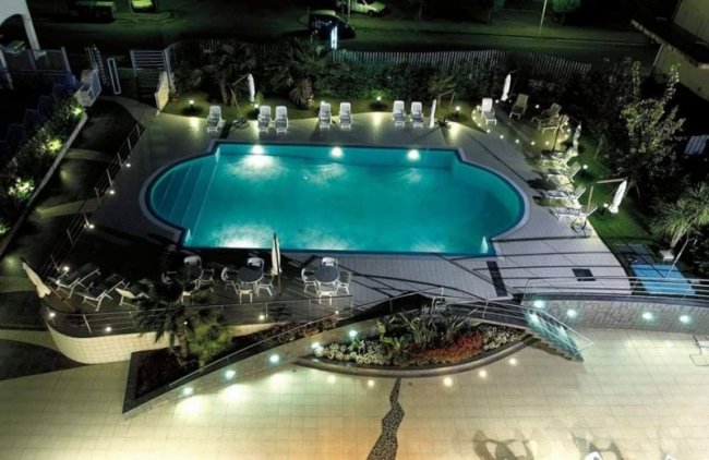 Piscina illuminata di notte con lettini e tavolini intorno.