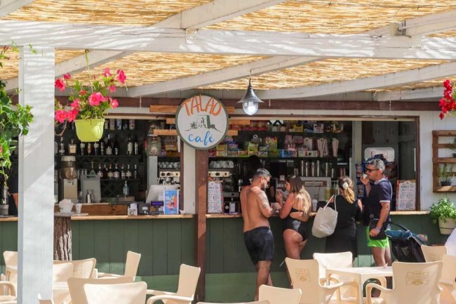 Un bar all'aperto con tetto di canne, persone al bancone e fiori appesi. Sedie di plastica beige.