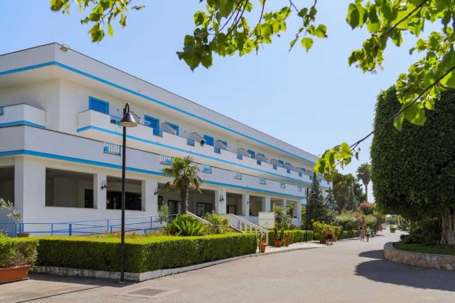 Edificio bianco a due piani con rifiniture blu, strada alberata e cespugli curati.
