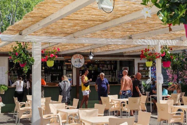 Bar all'aperto con tetto di canne, persone che socializzano, sedie beige e fiori appesi.