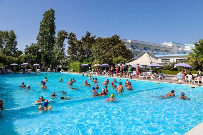 Persone in una grande piscina circondata da ombrelloni e alberi sotto un cielo sereno.