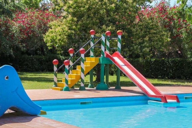 Uno scivolo colorato vicino a una piscina in un giardino alberato.