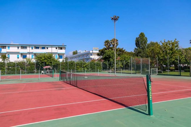 Un singolo campo da tennis con rete, circondato da alberi e edifici. Il cielo è sereno.