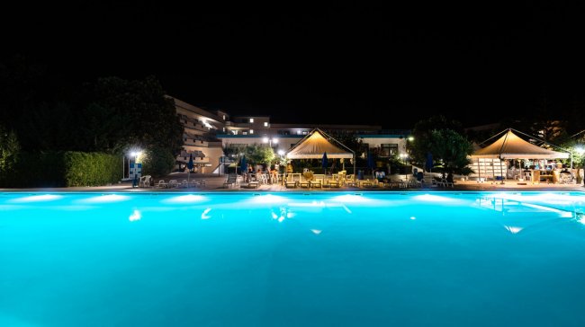 Una piscina illuminata di notte con strutture alberghiere sullo sfondo.