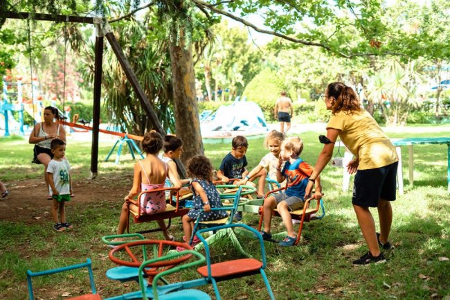 Bambini giocano su una giostra con l'aiuto di un adulto in un parco verde.