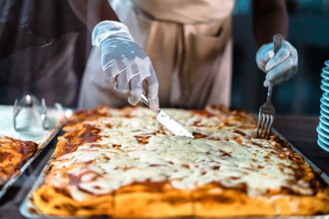 Mani con guanti affettano una pizza rettangolare su un vassoio.