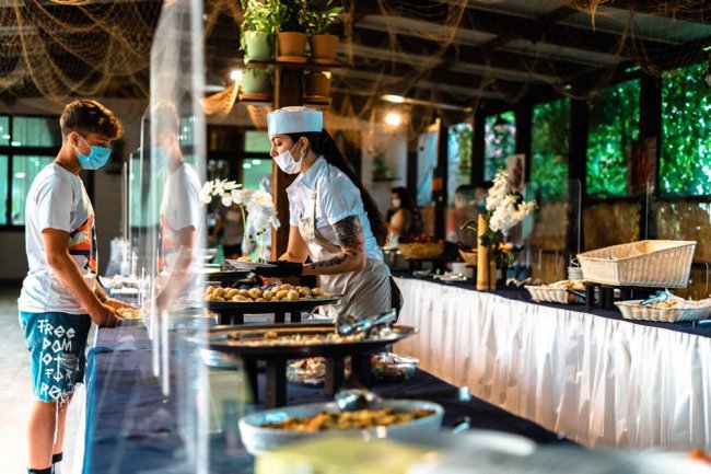 Buffet in un ristorante con una chef che serve un cliente, entrambi indossano mascherine.