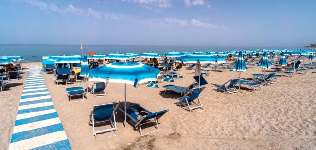 Spiaggia sabbiosa con ombrelloni e lettini blu, mare sullo sfondo.