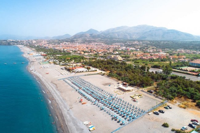 Veduta aerea di una lunga spiaggia con ombrelloni e una città sullo sfondo.
