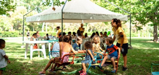 Bambini giocano in un parco sotto un gazebo, con adulti seduti ai tavoli.