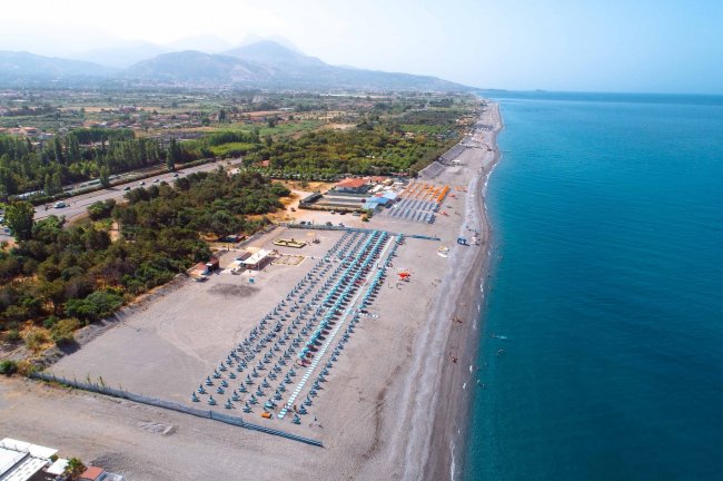Vista aerea di una lunga spiaggia con ombrelloni blu e mare calmo.