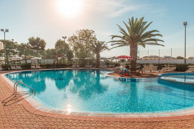 Piscina azzurra con palme e ombrelloni sotto il sole.