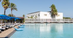 Piscina grande con lettini blu, palme sullo sfondo e un edificio bianco.