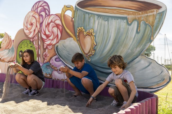 Bambini giocano con la sabbia davanti a tazze giganti.