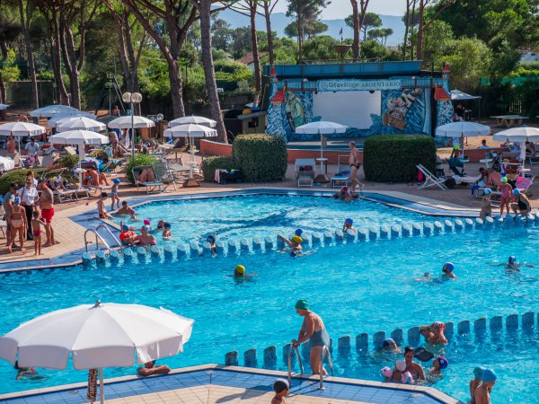 Piscina affollata con ombrelloni e scenografia sullo sfondo.