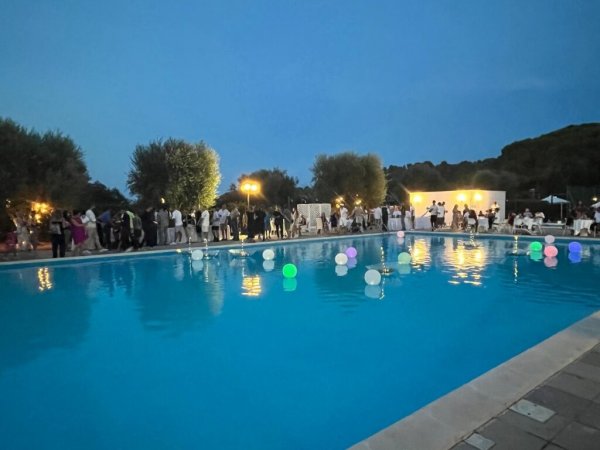 Piscina illuminata con palloncini galleggianti e persone sullo sfondo.