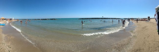 Spiaggia con persone in acqua vicino a barriere frangiflutti sotto un cielo limpido.