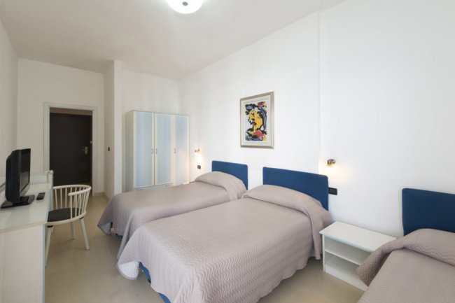 Stanza con tre letti singoli, coperte beige, armadio bianco e scrivania con sedia.