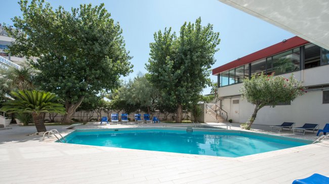 Una piscina circondata da lettini blu e alberi verdi, accanto a un edificio con pareti vetrate.