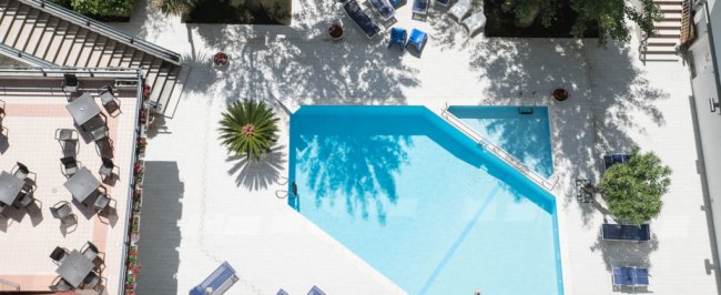 Piscina a forma irregolare vista dall'alto, circondata da lettini blu e palmizi.