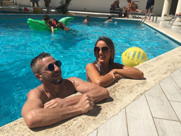 Due persone sorridenti al bordo di una piscina con un pallone giallo.
