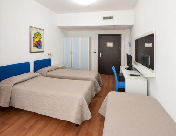 Stanza con tre letti singoli, armadio bianco e TV su scrivania. Quadro colorato appeso al muro.