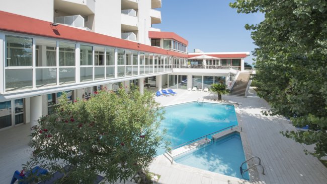 Piscina all'aperto circondata da un edificio bianco con balconi e vegetazione.