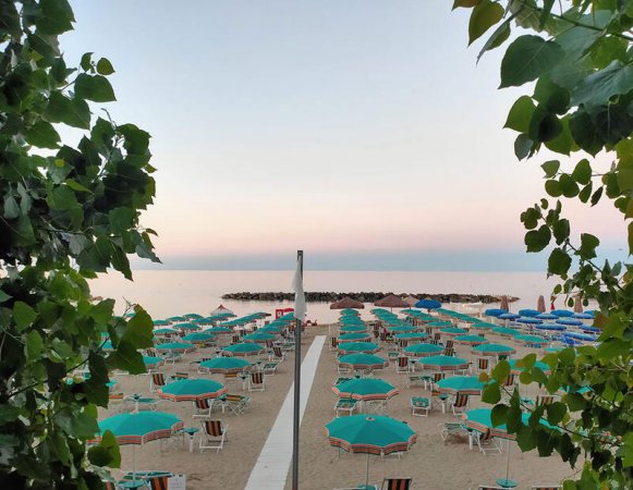 Spiaggia al tramonto con ombrelloni turchesi e mare calmo, vista tra alberi.