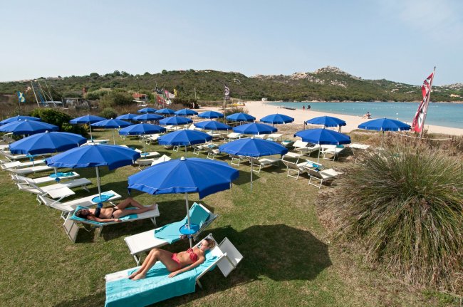 Lettini bianchi con ombrelloni blu su un prato verde vicino a una spiaggia.