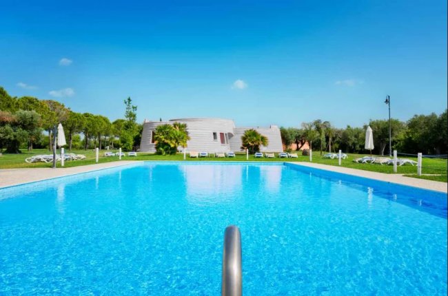 Piscina limpida all'aperto circondata da ombrelloni e prato verde, cielo blu.