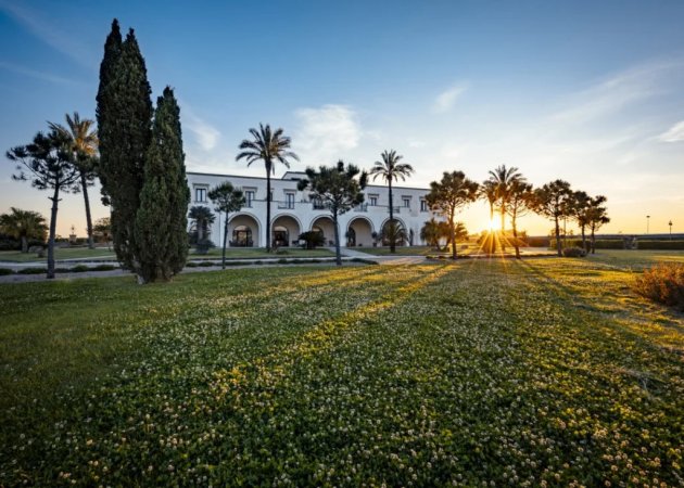 Edificio bianco con archi, alberi al tramonto su prato fiorito.