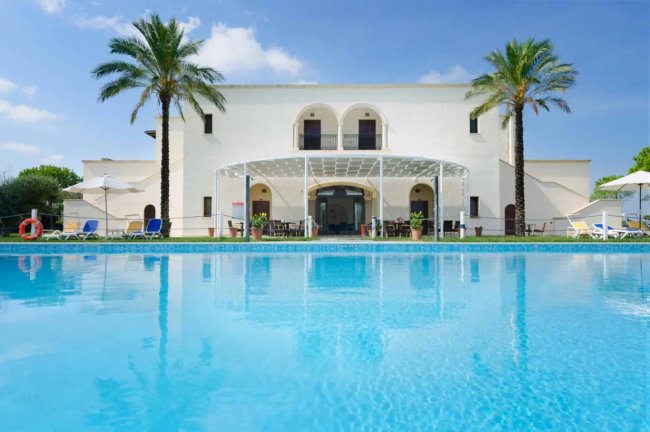 Grande villa bianca con piscina in primo piano e palme ai lati.