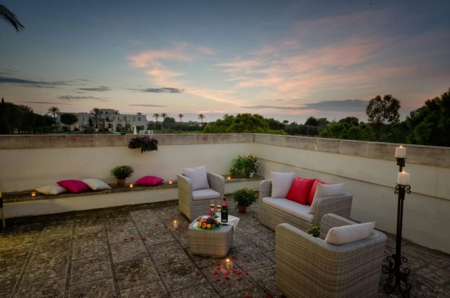 Terrazza con sedie di vimini, candele, cuscini colorati e bottiglia di vino al tramonto.