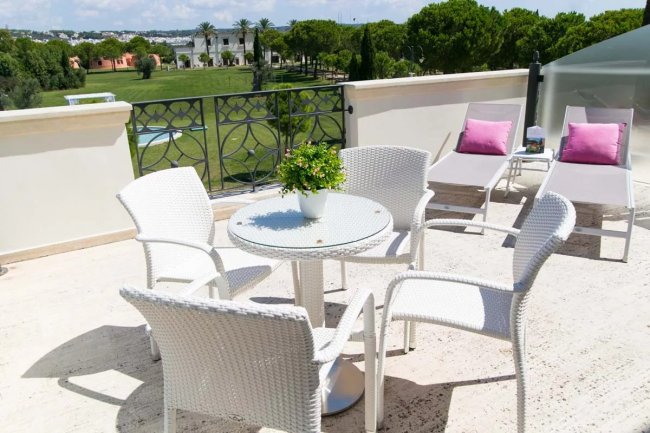 Terrazza con tavolino rotondo, sedie bianche, due lettini e cuscini rosa.