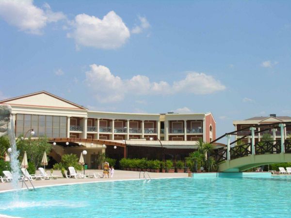Piscina esterna con ponte di legno e hotel sullo sfondo, cielo azzurro e poche nuvole.