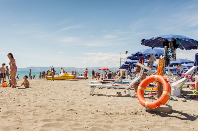 Spiaggia affollata con ombrelloni blu, sedie a sdraio e persone che si godono il sole e il mare.