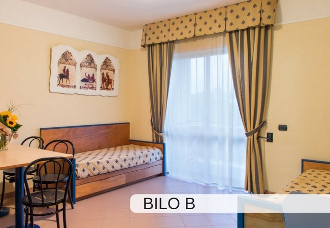 Camera d'hotel con due letti singoli, tavolo e sedie, tende e decorazioni a parete.