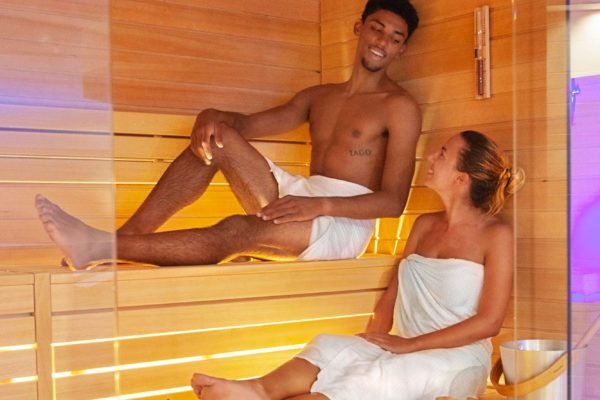 Due persone in una sauna, avvolte in asciugamani, che sorridono e conversano.