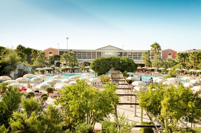 Giardino di un resort con piscine e ombrelloni, circondato da alberi e strutture alberghiere.