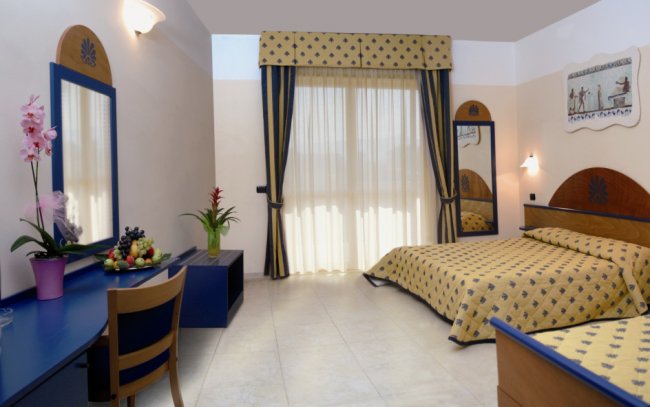 Camera da letto con letto matrimoniale, scrivania blu e grandi finestre con tende a righe.