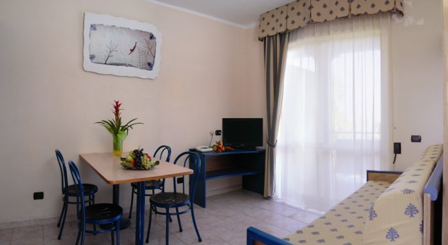 Soggiorno con tavolo, quattro sedie blu, piante e TV. Finestre con tende trasparenti.