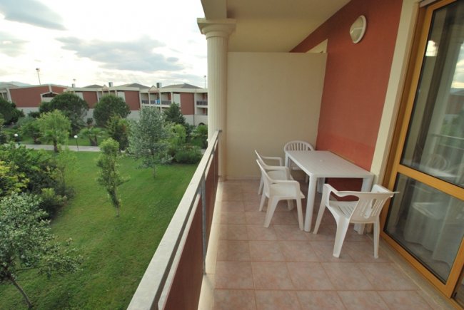 Balcone con tavolo e quattro sedie bianche, vista su un giardino e edifici.