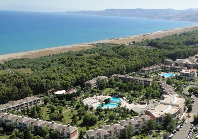 Vista aerea di un resort con piscine, giardini e la spiaggia. Mare e montagne sullo sfondo.